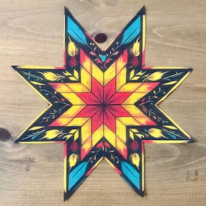 May include: A colorful star-shaped design with a geometric pattern. The star features a red center that transitions to yellow and is outlined in black. It is decorated with blue feathers, yellow leaves, and red pinecone-like shapes. The design is on a wooden surface.
