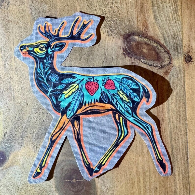 Op de afbeelding: Een kleurrijk geborduurd patch van een hert met een bloemen- en verenpatroon. Het hert is oranje omrand en heeft een blauw, groen en geel patroon. Er zitten twee rode aardbeien op de rug van het hert.