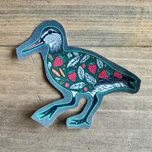 May include: A green and gray fabric patch of a bird with a long beak and legs. The bird is covered in a pattern of red strawberries, green leaves, and brown carrots.