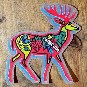 Op de afbeelding: Een rood hert silhouet met een blauwe omtrek, versierd met kleurrijke veren, aardbeien en bloemen. Het hert staat op een grijze achtergrond.