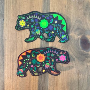 May include: Two black bear silhouette cutouts with colorful floral designs. The top bear has green, blue, and yellow flowers. The bottom bear has orange, pink, and red flowers.