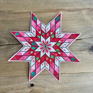 May include: A decorative star-shaped patch with a strawberry design. The patch features a geometric pattern of red, pink, and white sections, with strawberry and leaf illustrations. The star has a pink border and is set against a wooden background.