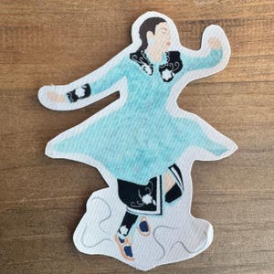 May include: A blue and black fabric patch depicting a woman in a traditional dance outfit. She is wearing a light blue dress with black trim and black leggings. She is holding her arms out to the side and her feet are in mid-air.