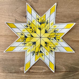 May include: A paper star with a yellow and white geometric design. The star's center is yellow with floral accents and dark blue leaves. The star is on a wooden surface.