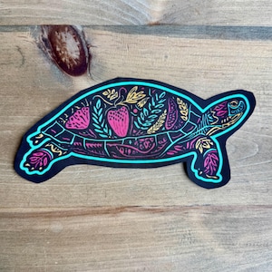 Peut inclure: Un autocollant découpé en forme de tortue avec un fond noir et un motif néon vibrant. La carapace présente des motifs botaniques complexes, incluant des feuilles et une fraise, dessinés avec des contours rose, jaune et cyan vifs.