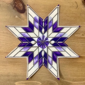 Peut inclure: Un objet décoratif en forme d'étoile avec un motif géométrique. L'étoile présente un motif de formes blanches et violettes, avec un motif central de lignes violettes rayonnantes. Les bords sont soulignés en noir, et l'effet global est complexe et détaillé.