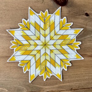 May include: A die-cut sticker featuring a starburst design with yellow and white geometric patterns. The star has yellow leaf accents around the edges. The sticker is on a wooden surface.