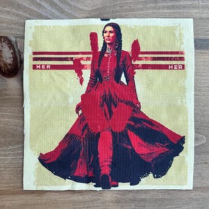 May include: A fabric square with a stylized illustration of a woman in a long red dress with a flowing skirt. The text "HER" is printed in red on a yellow background. The woman has long braids.