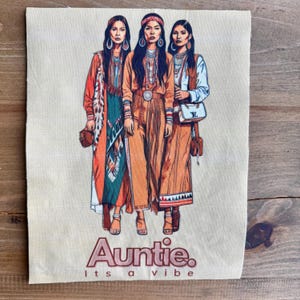 May include: Auntie graphic on a beige fabric. The illustration depicts three women in bohemian-style clothing, featuring long dresses and accessories. The text "Auntie. Its a vibe" is in a pink font at the bottom of the image.