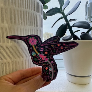 Peut inclure: Un colibri coloré avec des fleurs et des feuilles roses, bleues et vertes sur un fond noir. Le colibri est découpé et a une bordure blanche.