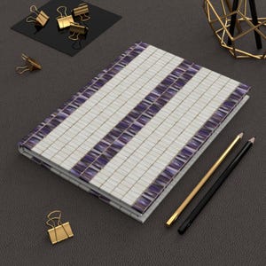 Pode incluir: Um caderno branco e roxo listrado com um design de mosaico. O caderno está sobre uma superfície cinza escura com dois lápis e um clipe de papel dourado.