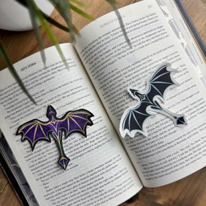 May include: Two black and white dragon stickers with purple and gold accents on an open book. The book has text about a fantasy story. The stickers are a great addition to any book lover's collection.
