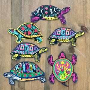 May include: Six colourful turtle stickers with detailed designs. The turtles display patterns of strawberries, feathers, and geometric shapes in vibrant colours such as pink, yellow, blue, and green. The stickers are arranged on a wooden surface.