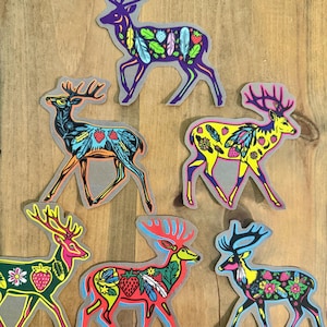 Op de afbeelding: Zes kleurrijke herten stickers met ingewikkelde ontwerpen met veren, aardbeien en bloemmotieven. De herten staan in verschillende poses en kleuren, waaronder paars, blauw, geel, rood en zwart.