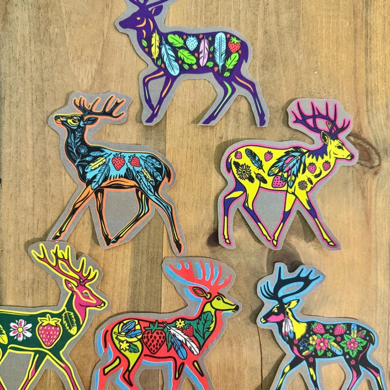 Op de afbeelding: Zes kleurrijke herten stickers met ingewikkelde ontwerpen met veren, aardbeien en bloemmotieven. De herten staan in verschillende poses en kleuren, waaronder paars, blauw, geel, rood en zwart.