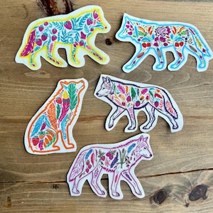 May include: A collection of five die-cut stickers featuring wolf designs. Each wolf is filled with colorful floral and berry patterns in shades of pink, blue, yellow, orange, and green. The stickers are on a wooden surface.