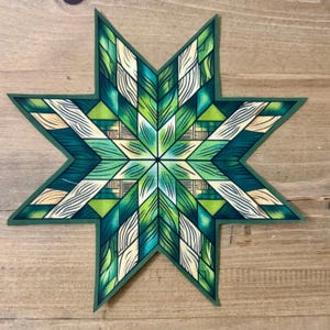 May include: A decorative star-shaped object with a geometric design. The star features shades of green, teal, and cream, with a dark green border. The design has a quilt-like appearance, with various shapes and patterns. The object is placed on a wooden surface.