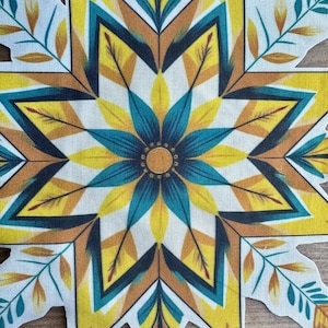 May include: A fabric cutout in the shape of an eight-pointed star featuring a central circular motif. The design displays a symmetrical pattern of yellow, teal, and brown leaf-like shapes radiating outward from the center on a white background.