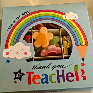 Puede incluir: Una caja de regalo azul con un dise&ntilde;o de arco&iacute;ris y nubes. La caja dice "You're the BEST!" y "Thank you Teacher" con una estrella y una A+ en la caja. La caja contiene una variedad de caramelos coloridos.