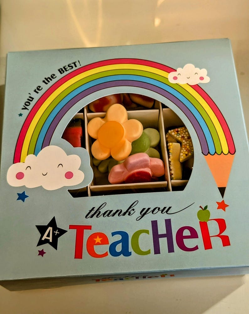 Puede incluir: Una caja de regalo azul con un dise&ntilde;o de arco&iacute;ris y nubes. La caja dice "You're the BEST!" y "Thank you Teacher" con una estrella y una A+ en la caja. La caja contiene una variedad de caramelos coloridos.
