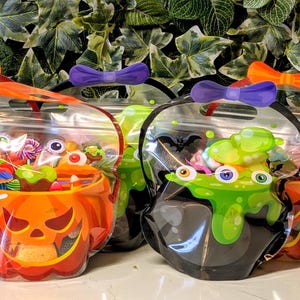 May include: Four clear plastic pouches with handles and bow-shaped closures. Two pouches feature an orange jack-o'-lantern print, and two feature a black cauldron print with green bubbling liquid and eyeballs, all filled with assorted sweets.