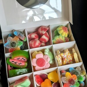 Puede incluir: Una caja blanca llena de caramelos gomosos de colores surtidos. Los caramelos est&aacute;n dispuestos en forma de cuadr&iacute;cula e incluyen rodajas de sand&iacute;a, flores, estrellas y corazones.