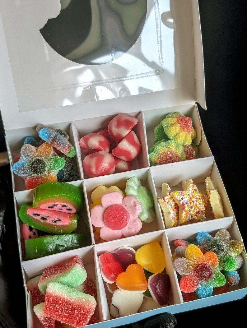 Puede incluir: Una caja blanca llena de caramelos gomosos de colores surtidos. Los caramelos est&aacute;n dispuestos en forma de cuadr&iacute;cula e incluyen rodajas de sand&iacute;a, flores, estrellas y corazones.