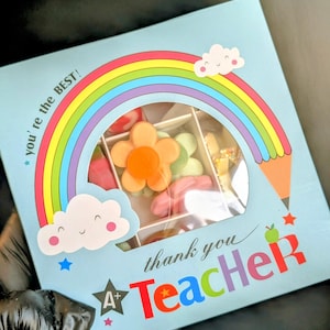 Puede incluir: Una caja de regalo azul con un dise&ntilde;o de arco&iacute;ris y nubes. La caja tiene una ventana que muestra una variedad de caramelos coloridos. El texto en la caja dice "you're the BEST!" y "thank you Teacher".