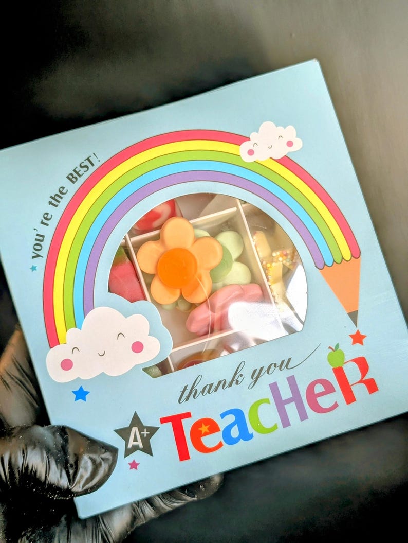 Puede incluir: Una caja de regalo azul con un dise&ntilde;o de arco&iacute;ris y nubes. La caja tiene una ventana que muestra una variedad de caramelos coloridos. El texto en la caja dice "you're the BEST!" y "thank you Teacher".