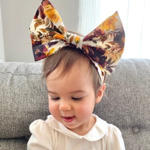 May include: A baby wearing a white collared shirt with a large floral print bow headband. The headband has a white background with burgundy, yellow, and orange flowers.