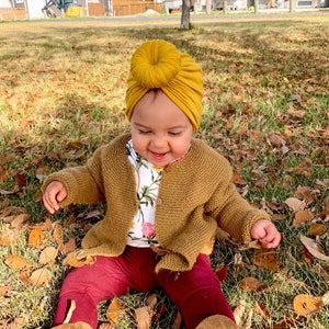 Puede incluir: Un niño pequeño con un turbante amarillo mostaza con un nudo en la parte superior, una chaqueta de punto marrón, leggings marrones y botas marrones. El niño está sentado en una zona cubierta de hierba con hojas caídas.