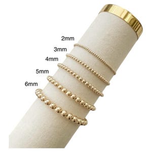 Könnte beinhalten: Goldene Perlenarmbänder in verschiedenen Größen, von 2 mm bis 6 mm, auf einem weißen zylindrischen Ständer präsentiert. Die Größen sind beschriftet. Die Armbänder sind goldfarben, mit gleichmäßig verteilten Perlen.