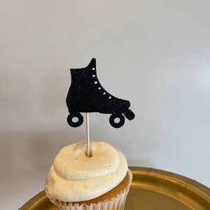 May include: A cupcake with white frosting and a black glitter roller skate topper. The cupcake sits on a gold plate. The roller skate is a silhouette with white dots for laces and wheels.