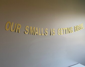 Può includere: Lettere dorate con glitter che compongono la scritta "OUR SMALLS IS GETTING BIGGIE" su una parete grigia.
