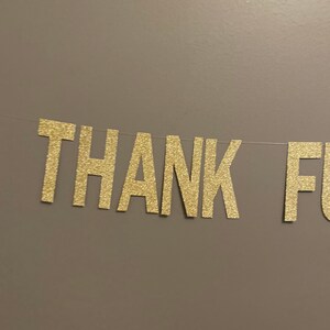 May include: Gold glitter letters spelling "THANK YOU" on a brown wall.