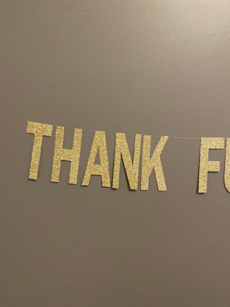 May include: Gold glitter letters spelling "THANK YOU" on a brown wall.