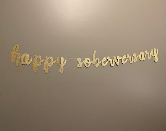 Może przedstawiać: Złota girlanda z błyskotkami z napisem "happy soberversary" wisząca na ścianie.