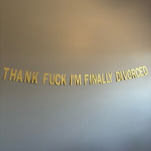 May include: A gold glitter banner with the text "THANK FUCK I'M FINALLY DIVORCED" hanging on a gray wall.