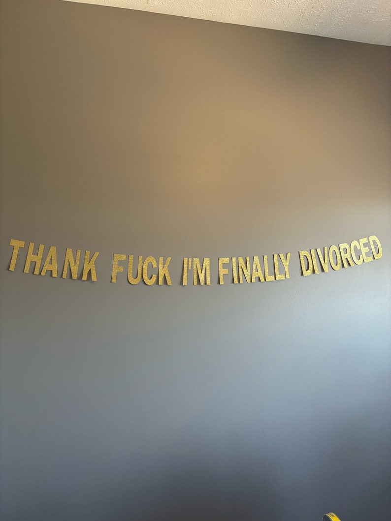 May include: A gold glitter banner with the text "THANK FUCK I'M FINALLY DIVORCED" hanging on a gray wall.