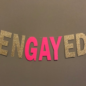 Könnte beinhalten: Goldene Glitzerbuchstaben, die "ENGAYED" buchstabieren, wobei das "GAY" in rosafarbenem Glitzer ist.