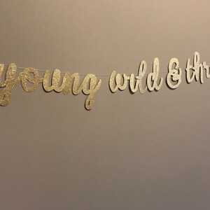 Puede incluir: Pancarta de purpurina dorada con el texto "young wild & three" colgada en una pared.
