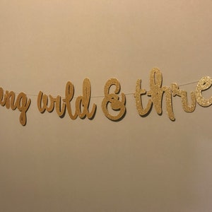 Puede incluir: Pancarta de purpurina dorada con el texto "young wild & three" para una fiesta de cumplea&ntilde;os.