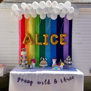 Puede incluir: Un tel&oacute;n de fondo de arco&iacute;ris con globos blancos que forman una forma de nube sobre las letras "ALICE" en globos de papel de aluminio dorado. Una mesa con un mantel blanco est&aacute; instalada frente al tel&oacute;n de fondo. La mesa est&aacute; decorada con cupcakes, figuras de unicornios y un letrero que dice "young wild & three".