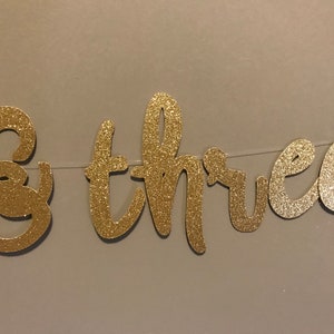 Puede incluir: Pancarta de brillo dorado con las palabras "& tres" para una fiesta de cumplea&ntilde;os.