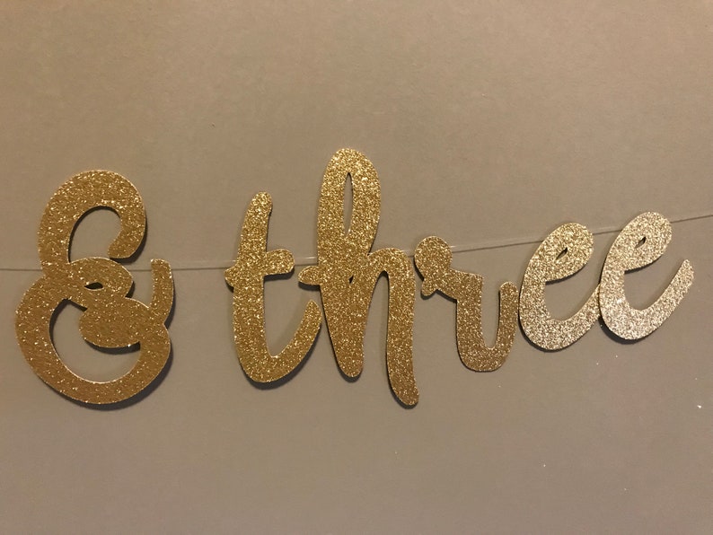 Puede incluir: Pancarta de brillo dorado con las palabras "& tres" para una fiesta de cumplea&ntilde;os.