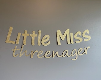 May include: Gold glitter letters spelling "Little Miss Threenager" on a gray background.