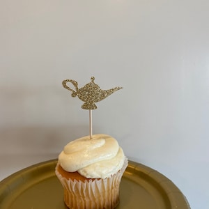 Puede incluir: Un cupcake blanco con glaseado de vainilla cubierto con un adorno de cupcake en forma de lámpara de genio de purpurina dorada. El cupcake está en un plato de papel dorado.