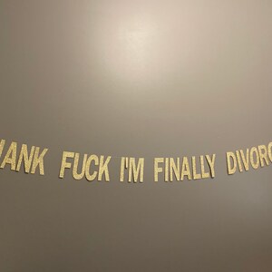 May include: Gold glitter letters spelling out "THANK FUCK I'M FINALLY DIVORCED" on a light brown wall.