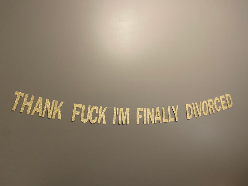 May include: Gold glitter letters spelling out "THANK FUCK I'M FINALLY DIVORCED" on a light brown wall.