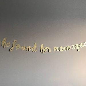 May include: Gold glitter letters spelling out the phrase "she found her own squeeze" on a gray wall.